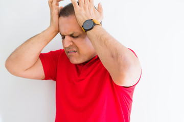 Naklejka premium Middle age man wearing red t-shirt over white wall suffering from headache desperate and stressed because pain and migraine. Hands on head.