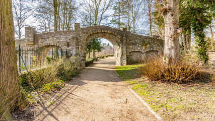 Old stone bridge with an arch over a dirt path leading to the Proosdij park, bare trees against a blue sky in background, sunny day in Meerssen, South Limburg in the Netherlands