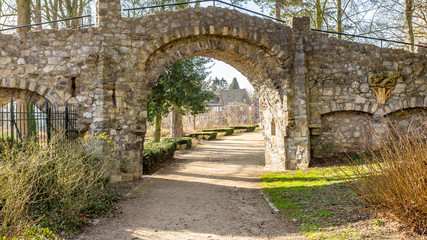 Fototapeta na wymiar Stone bridge with an arch with a dirt pedestrian path in the Proosdij Park with small bushes, trees and houses in the background, sunny winter day in Meerssen, South Limburg, Netherlands