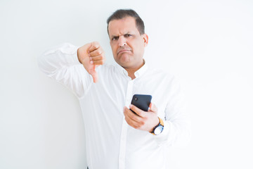 Middle age man using smartphone over white wall with angry face, negative sign showing dislike with thumbs down, rejection concept