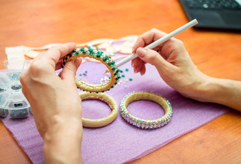 Close-up woman`s hands, decorating bracelets with colorful shiny rhinestones, gluing crystals on a textile base with wax pencil. Handicraft process concept. 