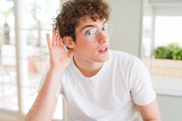 Young handsome man wearing glasses smiling with hand over ear listening an hearing to rumor or gossip. Deafness concept.