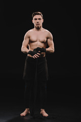 Fototapeta na wymiar strong shirtless and barefoot muscular mma fighter fixing bandages and looking at camera on black