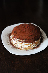 Freshly cooked tiramisu crepe cake dusted with powdery cocoa. White plate, wooden table