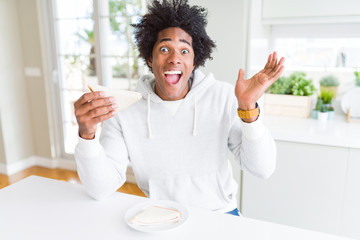Fototapeta na wymiar African American man eating handmade sandwich at home very happy and excited, winner expression celebrating victory screaming with big smile and raised hands