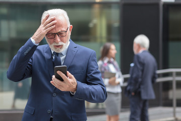 Worried senior gray hair businessman receiving bad news from business partners, holding his head. Two people talking in the background, blurred
