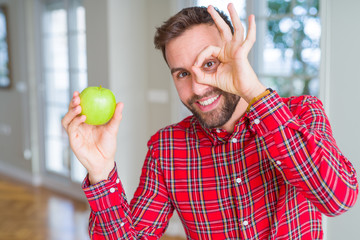 Handsome man eating fresh healthy green apple with happy face smiling doing ok sign with hand on eye looking through fingers