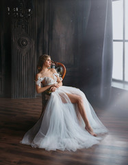Trendy woman bride in sexy white chic white silver bridal dress sitting on chair, dark room high...