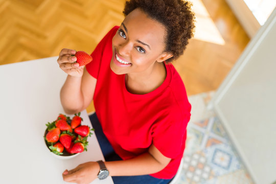 Beautiful young african american woman eating fresh red strawberries at home