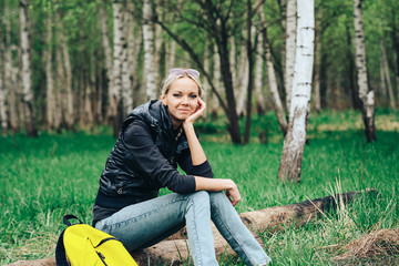 An attractive girl with a backpack on a walk in the Park sat down on a log and smiles in a good mood.