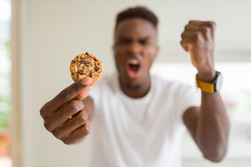 Young african american man eating chocolate chips cookies annoyed and frustrated shouting with anger, crazy and yelling with raised hand, anger concept