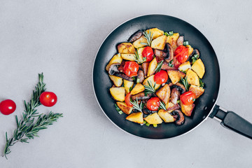Fried potatoes in a pan with tomatoes, mushrooms,greens decorated with sprigs rosemary on a  table vegetarian food Top view - Image