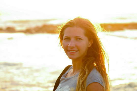 Candid portrait of young woman by the sea at sunset, strong sun backlight, background overexposed intentionally for atmosphere