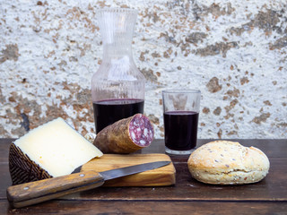 An Iberian pork sausage on an old wooden board with a wedge of sheep cheese, an antique knife and a bottle and a glass of red spanish wine