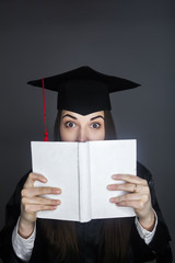 young graduate in a cap and prom dress looks out from behind the book