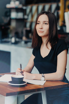 Pondering female student writes list to do in notepad, thinks on future plans, enjoys coffee break in cosy cafeteria, notes information with pen, concentrated into distance. People, leisure concept
