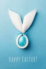 Easter bunny ears made of white napkin with egg on blue background. Minimal styled Happy Easter card concept.