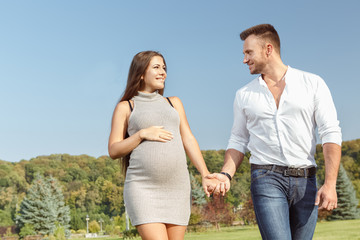 Fototapeta na wymiar Family walk. Cheerful pregnant couple walking in the park holding hands and smiling happily