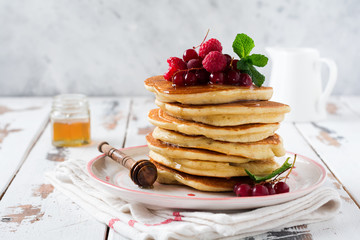Stack of homemade little pancakes with honey, fresh raspberries and red currants on an old  light wooden background. Selective focus.