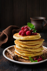 Stack of homemade little pancakes with honey, fresh raspberries and red currants on an old wooden background. Selective focus.