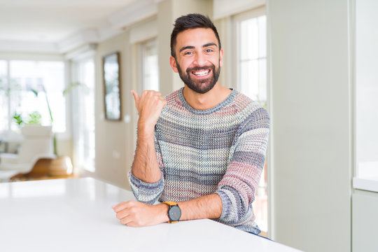 Handsome hispanic man wearing casual sweater at home smiling with happy face looking and pointing to the side with thumb up.