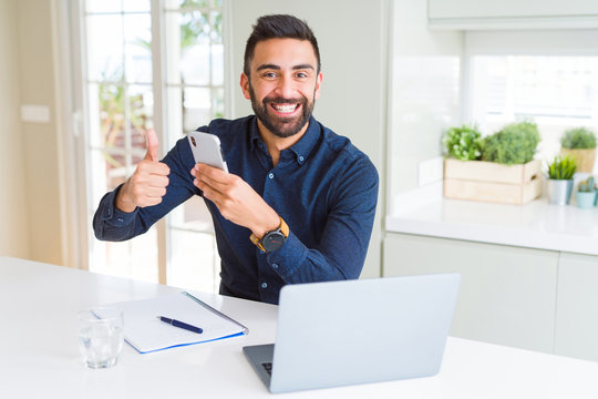 Handsome hispanic business man using smartphone and laptop at the office happy with big smile doing ok sign, thumb up with fingers, excellent sign