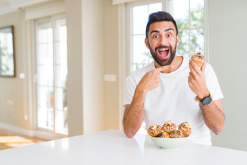 Handsome hispanic man eating chocolate chips muffin very happy pointing with hand and finger