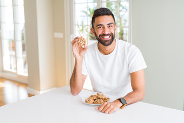 Handsome hispanic man eating chocolate chips cookies with a happy face standing and smiling with a confident smile showing teeth