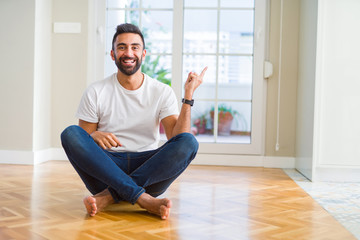 Handsome hispanic man wearing casual t-shirt sitting on the floor at home with a big smile on face, pointing with hand and finger to the side looking at the camera.