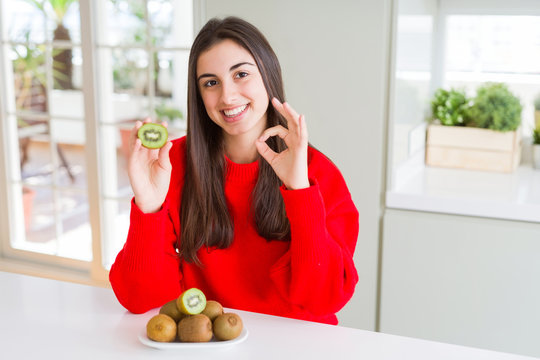 Beautiful young woman eating half fresh green kiwi doing ok sign with fingers, excellent symbol
