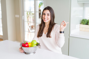 Beautiful young woman using colander to wash and clean vegatables very happy pointing with hand and finger to the side