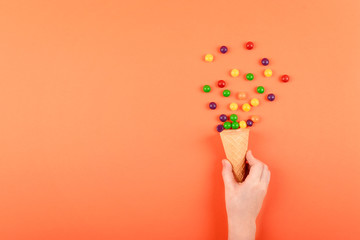 Child hand with waffle cone and sweet multicolored dragee on coral background. Festive, birthday, Easter composition, greeting card