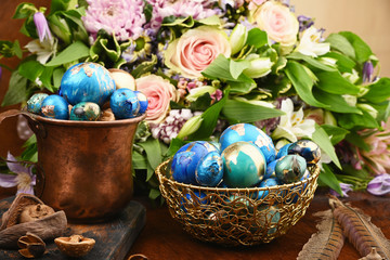 Painted colored Easter eggs in golden bowl and copper mug near bouquet of flowers on red animal skin. Boho stile.
