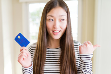 Beautiful Asian woman holding credit card pointing and showing with thumb up to the side with happy face smiling