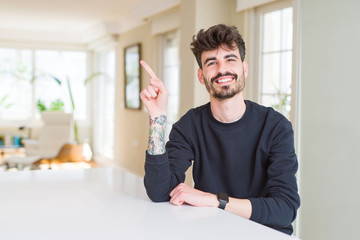 Young man wearing casual sweatshirt sitting on white table with a big smile on face, pointing with hand and finger to the side looking at the camera.