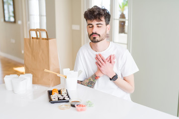 Young man eating asian sushi from home delivery smiling with hands on chest with closed eyes and grateful gesture on face. Health concept.