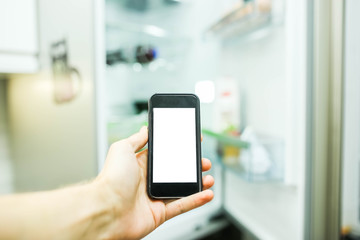 Smartphone with a white screen on the blurred background of an open refrigerator with products in the kitchen. Smart home technology system.