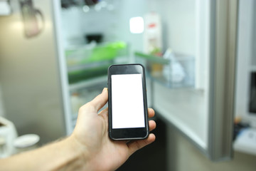 Smartphone with a white screen on the blurred background of an open refrigerator with products in the kitchen. Smart home technology system.