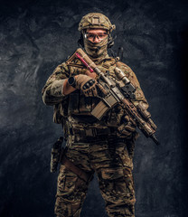 Fully equipped soldier in camouflage uniform holding an assault rifle. Studio photo against a dark textured wall