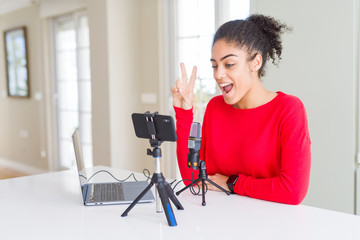 Young african american woman doing video call using smartphone camera and microphone smiling with happy face winking at the camera doing victory sign. Number two.