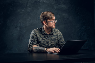 Programmer wearing a trendy military shirt sitting at the table with laptop in office against a dark wall, looking sideways with a thoughtful look - obrazy, fototapety, plakaty