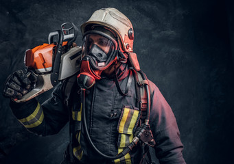 A brave firefighter wearing full protective equipment posing with a chainsaw on his shoulder. Studio photo against a dark textured wall
