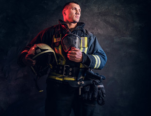 Fototapeta na wymiar Brutal firefighter looks sideways and holds a helmet in his hand in the studio on a black background