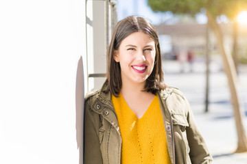 Beautiful young woman smiling confident and cheerful leaning on the wall, walking on the street of the city on a sunny day