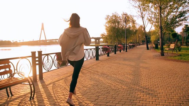 back view woman wearing grey hoodie and black leggings jogging along bank river with blue water female doing cardio workout near house