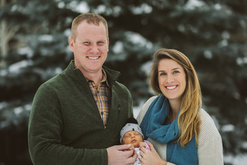 new mom and dad hold hand of newborn baby girl outside looking at camera smiling