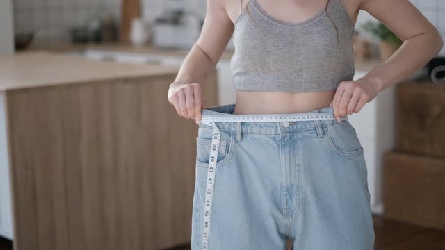 Concept of a healthy lifestyle. Young girl lost weight before summer season. Beautiful woman in bedroom measures waist with white measuring tape. Female with sport figure after successful diet