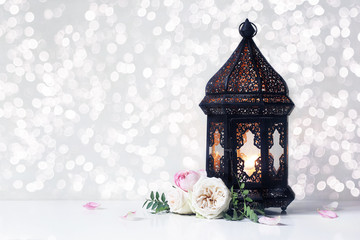 Fototapeta premium Vintage black Moroccan, Arabic lantern with glowing candle, green branches, rose flowers and petals on white table background. Greeting card for Muslim holiday Ramadan Kareem with bokeh lights.