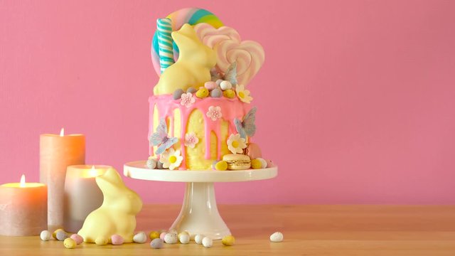 On trend Easter candy land drip cake decorated with lollipops, cand eggs and white chocolate bunny in pink party table setting.