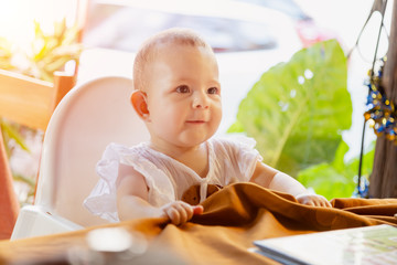 Fototapeta premium Infant girl is sitting on a baby high chair in a street cafe by the table. Children in white dress dabbles and pulls the tablecloth
